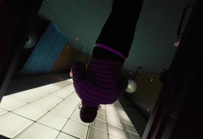 A person in a purple jacket hangs upside down in a dark room with a bright gridlike floor suggesting a dramatic surreal or scifi environment the camera is angled to emphasize the inverted perspective and the glowing floor panels