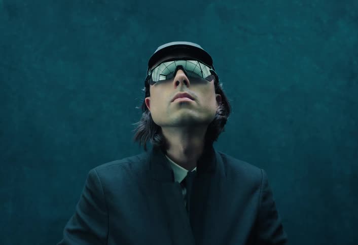 Person poses with mirrored sunglasses and dark clothing against a blue background