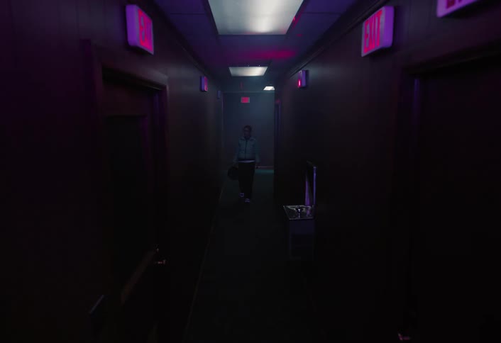 A dim hallway glows with pink and blue exit lights as a female walks at the far end creating a tense eerie vibe