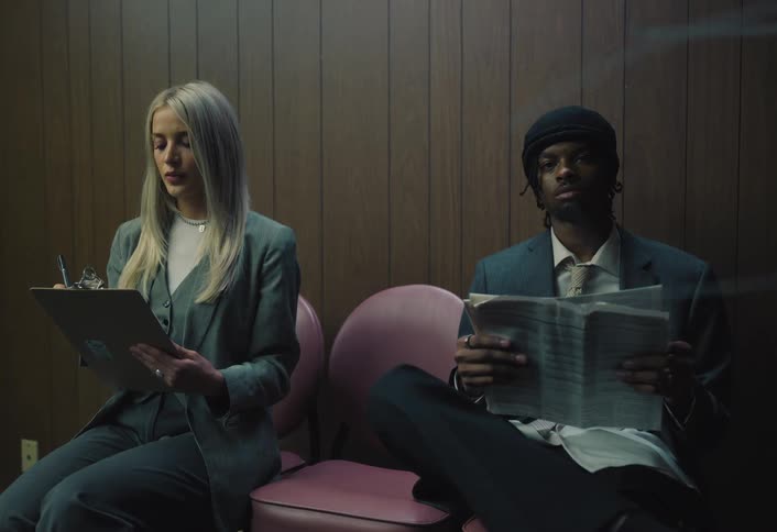 Two professionals sit waiting one reading a newspaper and the other holding a clipboard