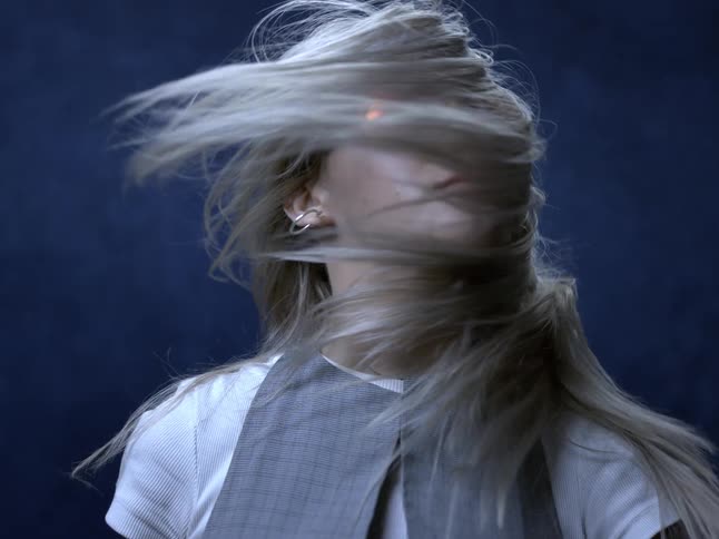 The video clip is a closeup midshot of a woman whose long lightcolored hair is dramatically blowing around her head she is wearing sunglasses and jewelry and her hair is whipping wildly against a dark blue background