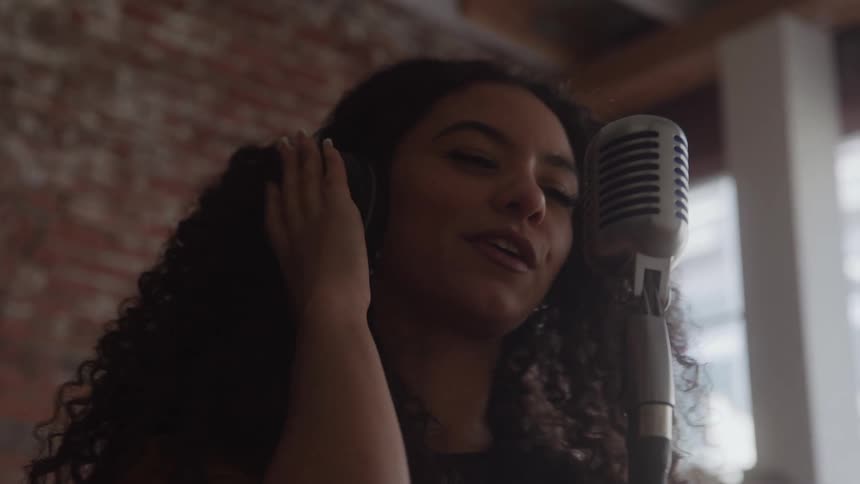 This is a tight closeup shot of a woman with long curly hair singing soulfully into a large vintage microphone she is closing her eyes and touching her ear with a softfocus brick wall in the background