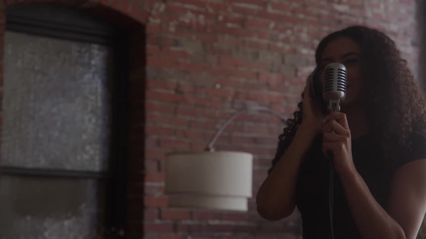 The video is a mediumcloseup shot of a woman with long curly hair singing passionately into a vintage microphone in an indoor setting the background features an exposed brick wall and a dark window or door frame on the left