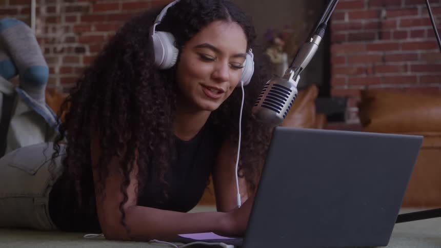 The face of a young girl with curly hair wearing headphones and speaking into a microphone in front of a laptop