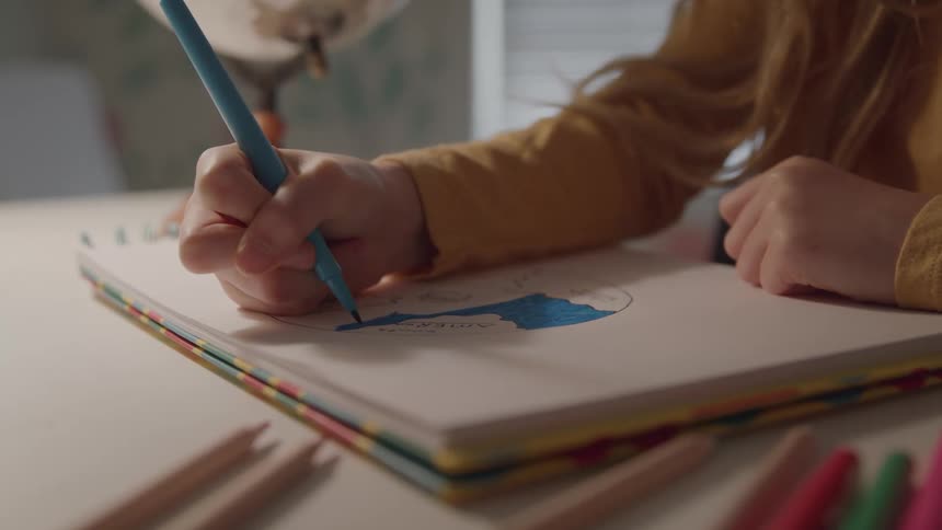 A young girl sits at a desk and colors a drawing in her notebook with a marker