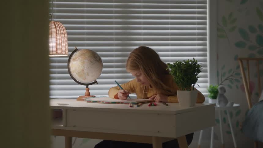 This is a wide shot looking through a doorway showing a young girl sitting at a white desk in a bedroom intently drawing or coloring in a notebook the camera slowly zooms out and reveals a wickershaded lamp and a globe on the desk with light filteri