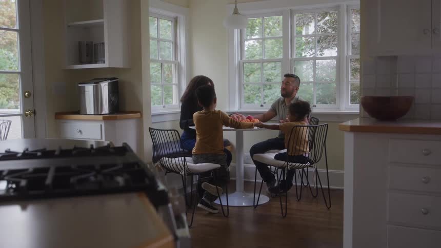 Family holds hands while praying at the breakfast table near their kitchen