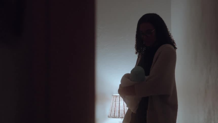 The video is a dimly lit medium shot of a woman in an apartment hallway seen in silhouette as she gently holds and cradles a bundled baby a wickershaded lamp provides a warm light source on a side table to the right illuminating her from behind
