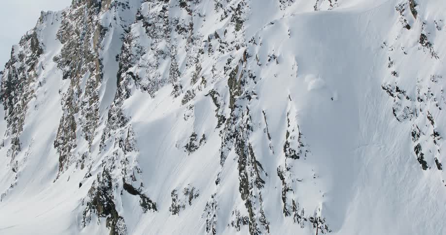 A vertical expanse of a mountain face covered in a mix of white snow and dark rock crags is shown with a person skiing down the snowcovered slope on the right side