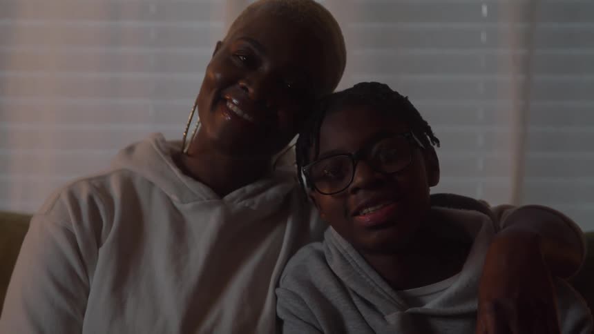 The video is a mediumcloseup shot of a mother and son sitting close together smiling at the camera in a dimly lit home interior the woman has short blonde hair and wears large hoop earrings and both are dressed in hooded sweatshirts