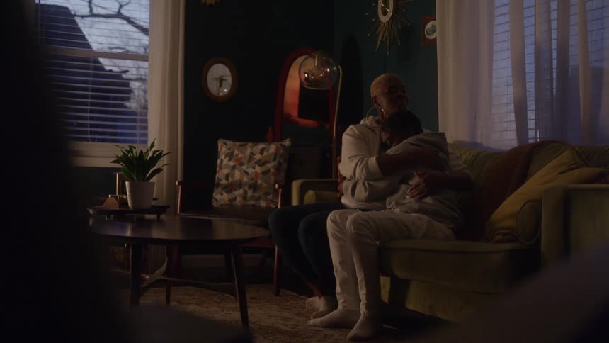 The video is a wide dimly lit shot of two people in lightcolored hoodies and pants sitting close together on a green sofa in a retrostyled living room the person on the left is embracing the person on the right in a tender comforting manner