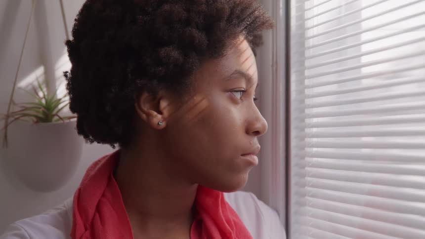 The video is a mediumcloseup profile shot of a young black woman with natural dark tightly curled hair she is seen looking out of a window partially covered by horizontal blinds with a serious or pensive expression