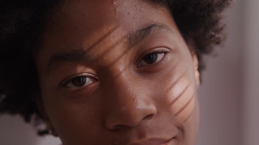 A closeup shot of a womans face shows parallel lines of shadow cast across the eyes and cheeks as the person looks directly toward the camera