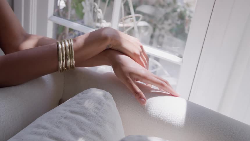 Closeup of a womans hands with bangles resting on a couch by a sunlit window