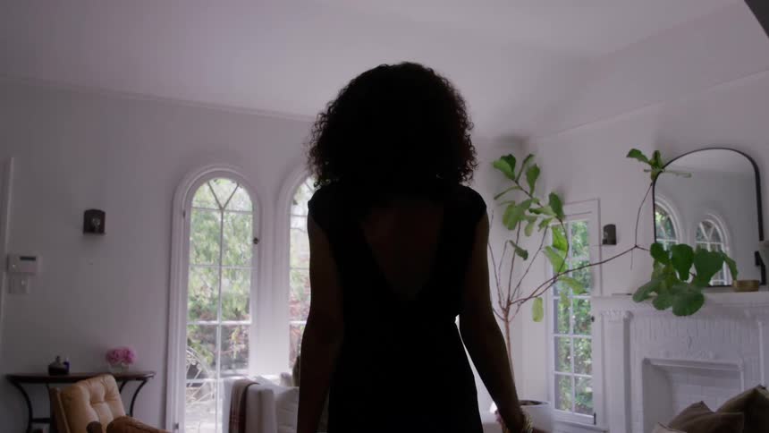 Woman wearing a fashionable low back black dress flips her hair while walking into a sunny living room