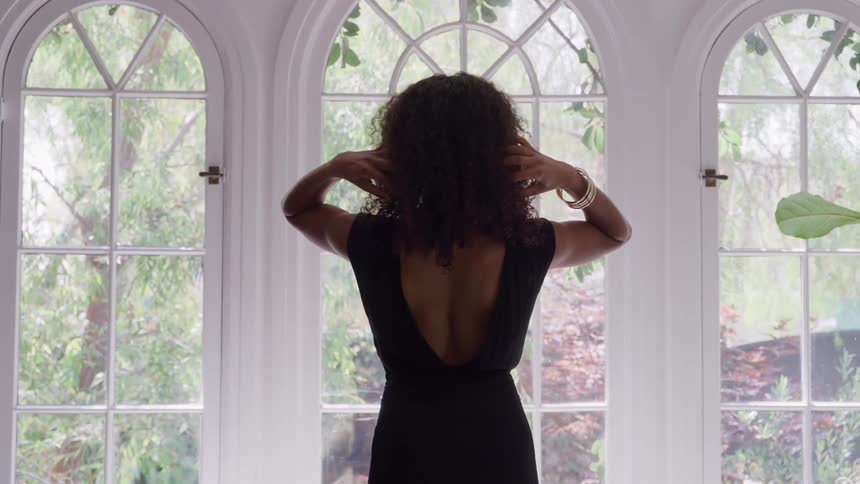 Woman stands adjusting her hair in a sunlit living room by a large window