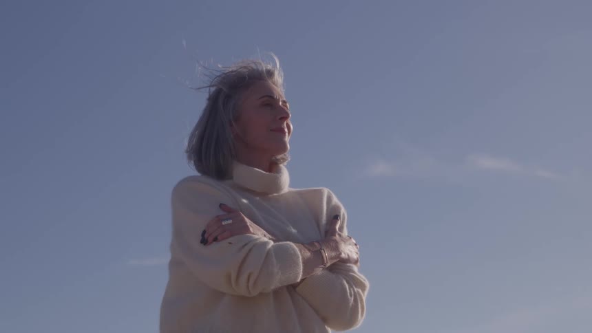 The video is a mediumwide profile shot of an older woman with gray hair standing outdoors against a light blue sky she is wearing a lightcolored turtleneck sweater and is wrapping her arms around herself with her hair gently blowing in the win