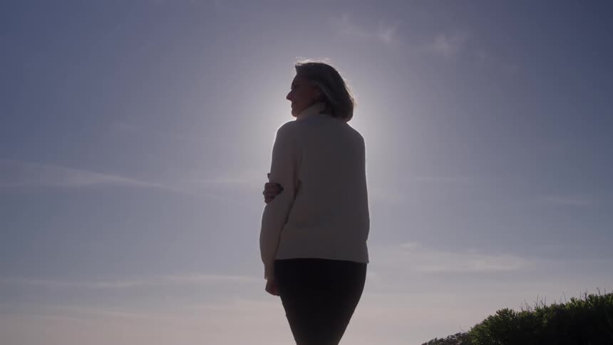 Woman stands with arm across her chest silhouetted against the sky outdoors