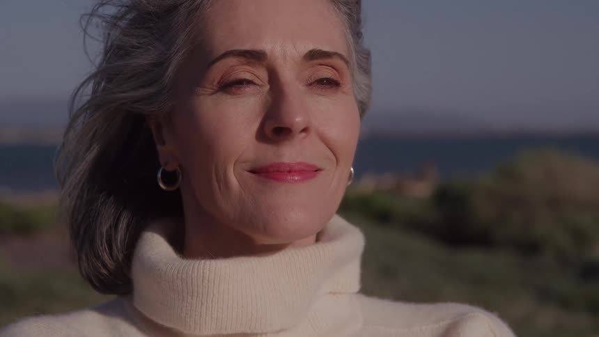 The video is a mediumcloseup profile shot of an older woman with gray hair standing outdoors on a sunny breezy day she is wearing a light turtleneck sweater and a silver hoop earring and she closes her eyes briefly before opening them with a calm co