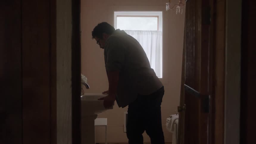The video shows a mediumwide shot of a man standing in a shadowy doorway leaning over a sink in a brightly lit bathroom he is wearing glasses and a collared shirt and is framed by the dark wood of the rooms entrance