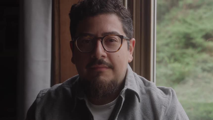 Man sits by a large window looking directly into the camera indoors