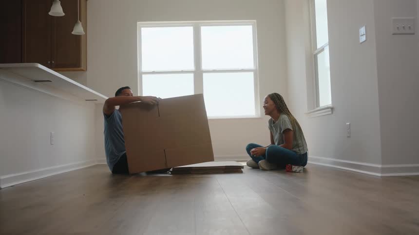 A man and a woman sit crosslegged on a wooden floor in a bright empty room near a large window with the man holding up a section of a cardboard box