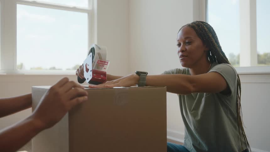 A woman smiles as she and another person tape up a cardboard box