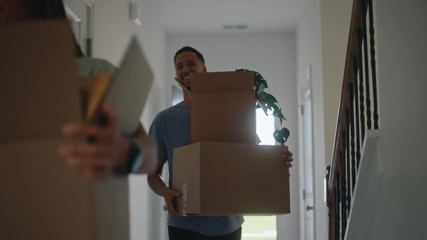 A smiling couple carries moving boxes into a bright hallway looking excited to be in their new home