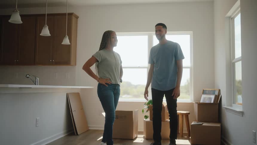 A man and a woman stand in the center of a newly occupied room with white walls and wood flooring facing the camera and smiling with several cardboard boxes near a large window