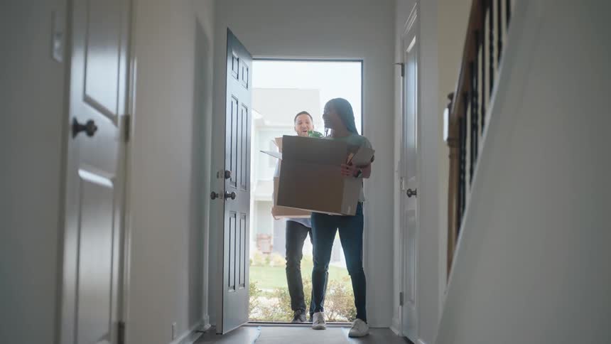 The video is a lowangle wide shot of a young woman entering a home through the front door carrying a large cardboard box she is smiling and wearing blue jeans and the bright view outside suggests it is a sunny day as she moves in or receives a delive
