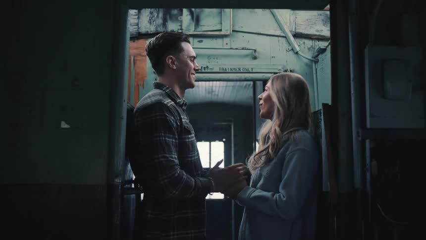 The video is a lowangle mediumcloseup shot of a young couple standing and holding hands in a dimly lit narrow passage possibly inside a train or old carriage they are facing each other and smiling with the man wearing a plaid shirt and the woman w