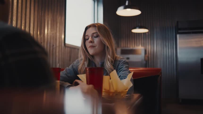 The video clip focuses on a young woman sitting at a booth in a dimly lit diner or restaurantshe is looking across the table at someone whose back is partially visible in the foreground with a serious or pensive expression