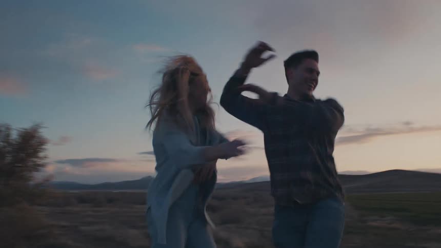 A man and a woman dancing in an open field with hills in the background under a fading sky facing each other and smiling