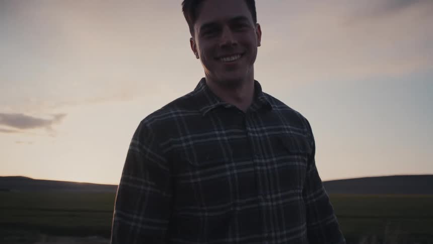 The video is a mediumcloseup shot of a young man with dark hair wearing a plaid buttonup shirt smiling directly at the camera he is standing outdoors at dusk or sunrise in a vast dry landscape of scrub and mountains