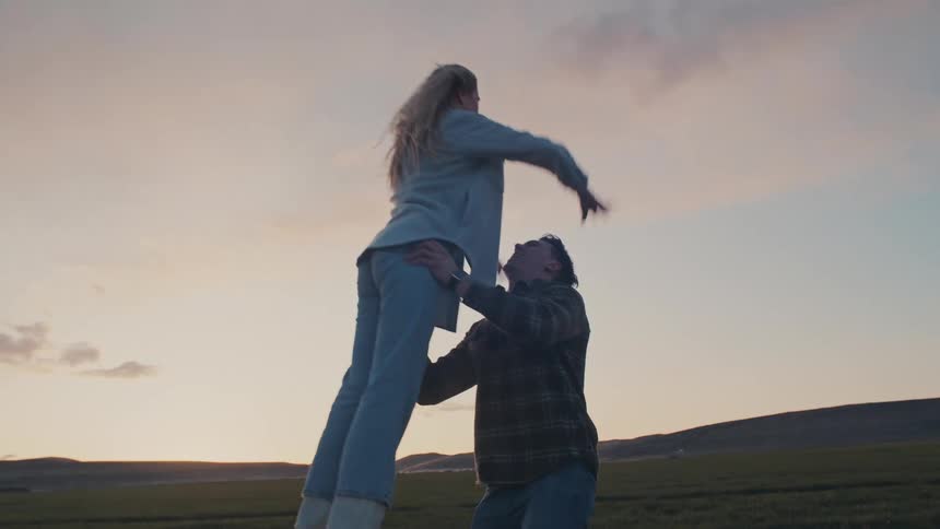 A man and a woman stand in a field with flat terrain in the background under a pale sky where the woman jumps up and is lifted by the man before they share a close embrace and smile at each other