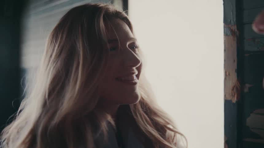 A woman with long hair and a man stand facetoface in a dimly lit space as the man reaches up and gently touches the womans cheek and they look into each others eyes and smile