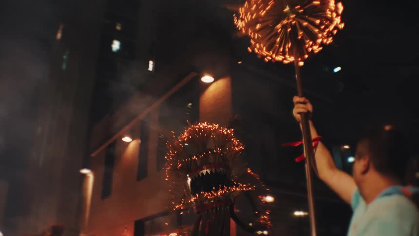 Multiple people carry glowing ceremonial objects down a dark city street at night with the crowd taking photographs in the foreground