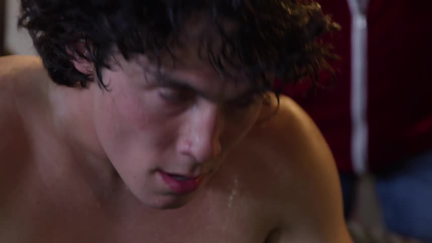 A male wearing green boxing gloves is sitting and breathing heavily as a person stands next to him