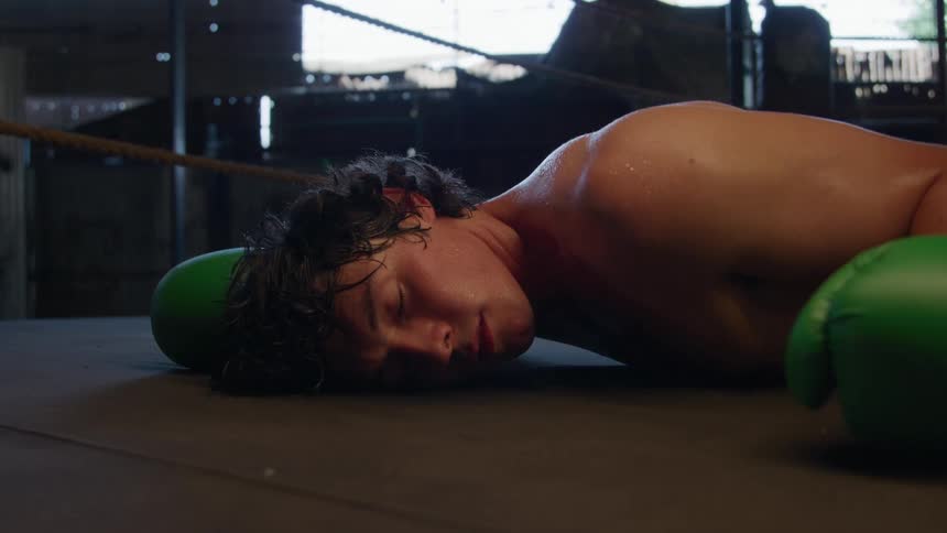 A young male boxer lies on the ring floor after his head falls to the mat between green gloves appearing dazed and exhausted