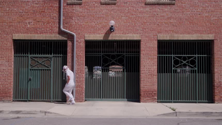 A person wearing all white clothing dances from right to left along a concrete sidewalk in front of a red brick building facade with three large grated metal doors