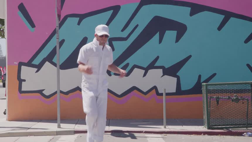 A person wearing all white clothing including a cap and sunglasses performs a dancing movement in front of a wall covered with bright teal and black graffiti on an urban sidewalk