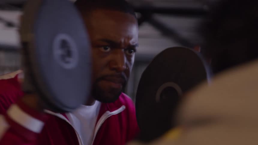 A man in a red track jacket and gloves holds two round focus pads out in front of him receiving punches from an offcamera person while looking on with an intense expression of concentration and effort