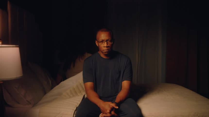 The video clip features a medium shot of a man wearing glasses and a dark tshirt sitting on the edge of a bed in a dimly lit bedroomhe looks directly at the camera with a solemn or troubled expression and remains motionless for the duration of the cl