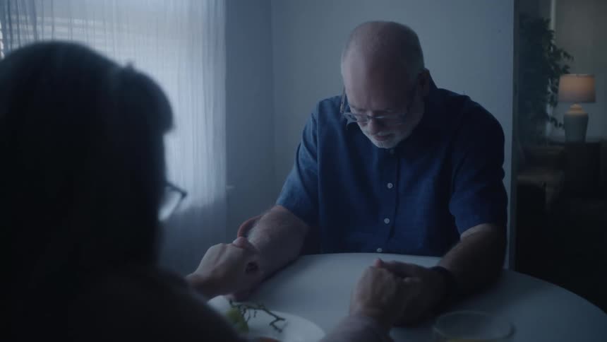 A couple holds hands and prays over a table with fruits indoors