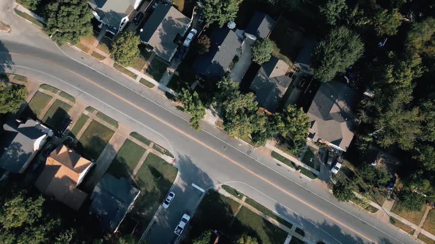 The video is an overhead drone shot of a diagonal stretch of a suburban street lined with houses and green treesthe street has a double yellow center line and parallel parking spaces along the curb highlighting the residential neighborhood lay