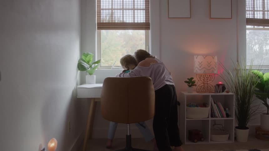 Two girls in a softly lit room one sitting at the desk and the other leaning in for a gentle hug surrounded by plants and warm light