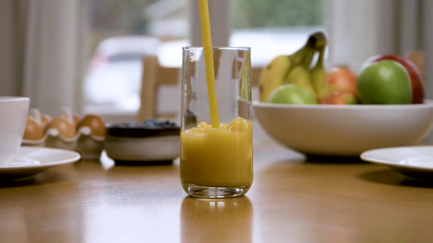 A stream of orange liquid is poured from above into a tall clear glass positioned in the center of a wooden table which is also set with cups a bowl of fruit and an egg carton in front of a bright window