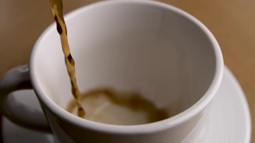 Closeup of dark coffee droplets falling into an empty white ceramic cup and saucer on a warm wooden surface