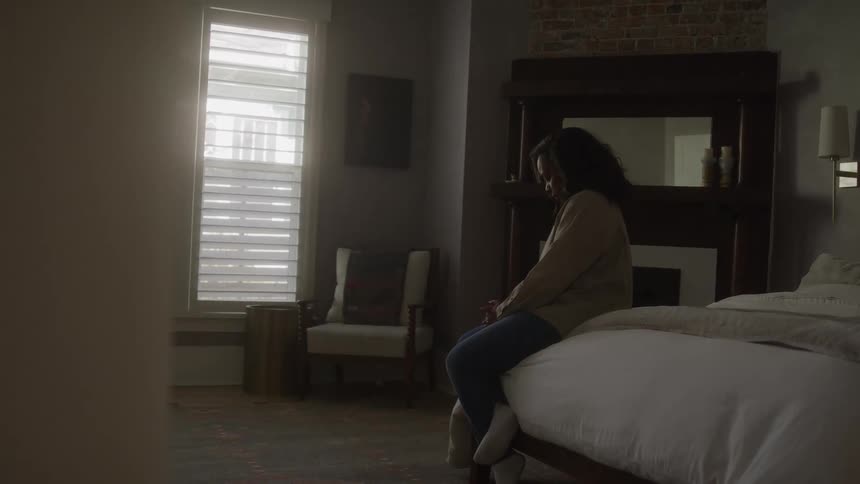 A person in a light shirt and dark pants sits on the edge of a bed in a dimly lit room facing away from the camera towards two tall windows with closed shutters while natural light illuminates the room