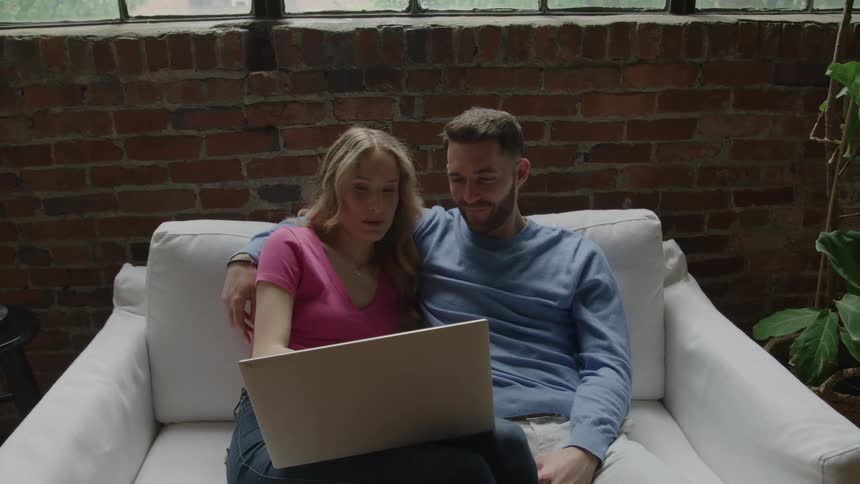 Couple sitting on a white sofa smiling while looking at a laptop